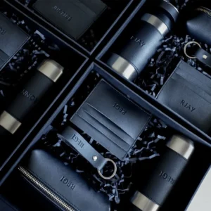 Prabangios verslo dovanos vyrams: odiniai raktų pakabukai, dėklai ir gertuvės su įspaustais inicialais juodose dėžutėse.