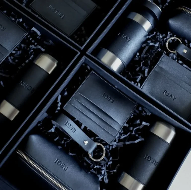 Prabangios verslo dovanos vyrams: odiniai raktų pakabukai, dėklai ir gertuvės su įspaustais inicialais juodose dėžutėse.