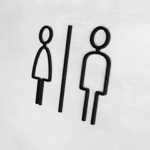 Modernūs, išpjauti juodo akrilo tualeto simboliai – vyras ir moteris – pritvirtinti tiesiai ant šviesios tekstūrinės sienos.