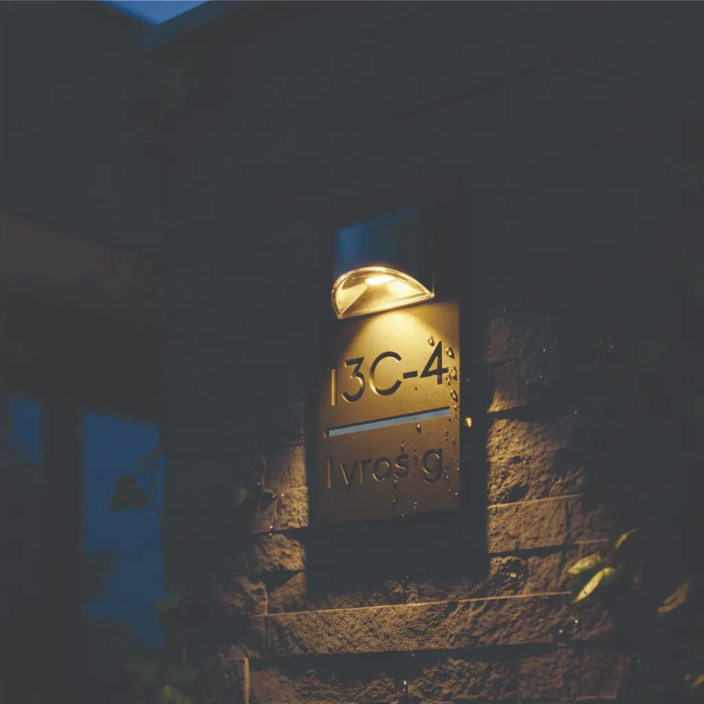 Moderni, apšviesta juodo ir veidrodinio akrilo adreso lentelė su užrašu „Lyros g 13C-4“ ant akmeninės fasado sienos vakaro metu.