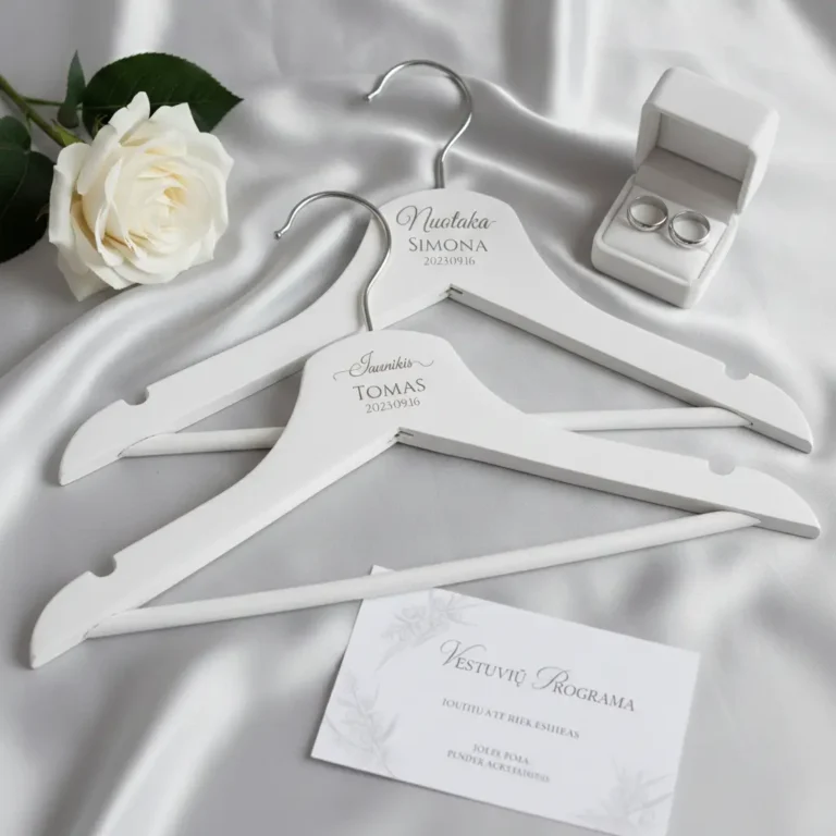 Personalizuota vestuvinė atributika: baltos matines pakabos su lazeriu graviruotais vardais, vestuviniai žiedai ir programa ant balto šilko.
