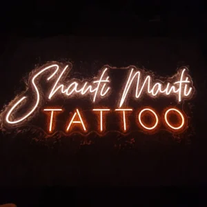 Rankų darbo LED neoninis užrašas „Shanti Manti TATTOO“ su elegantišku šviesiai gelsvu šriftu ir ryškia oranžine spalva tatuiruočių studijos interjerui.