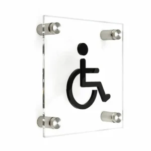 Skaidraus akrilo neįgaliųjų vežimėlio simbolio lentelė su keturiais nerūdijančio plieno distanciniais laikikliais ant baltos sienos.