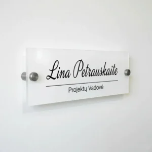 Elegantiška vardinė biuro lentelė ant sienos iš balto akrilo su juodu užrašu „Lina Petrauskaitė – Projektų Vadovė“ ir metaliniais laikikliais.