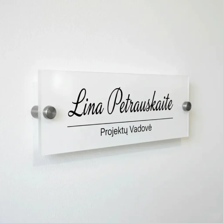 Elegantiška vardinė biuro lentelė ant sienos iš balto akrilo su juodu užrašu „Lina Petrauskaitė – Projektų Vadovė“ ir metaliniais laikikliais.