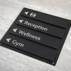 Matine juoda modulinė informacinė lentelė su baltais užrašais Reception, Wellness, Gym ir piktogramomis ant akmens tekstūros fono