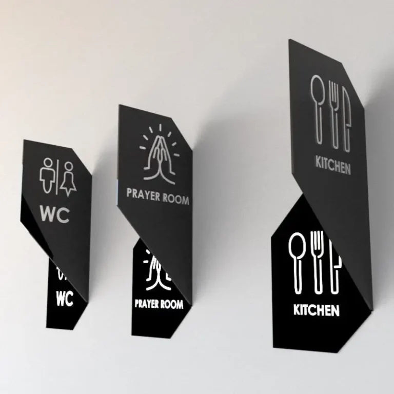 Tamsiai pilkų kampinių nuorodų rinkinys ant sienos su baltais simboliais ir užrašais: WC, Prayer Room (maldos kambarys) ir Kitchen (virtuvė)