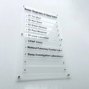 Skaidraus akrilo modulinė informacinė lentelė su juodais užrašais ir metaliniais distanciniais laikikliais ant šviesios sienos.